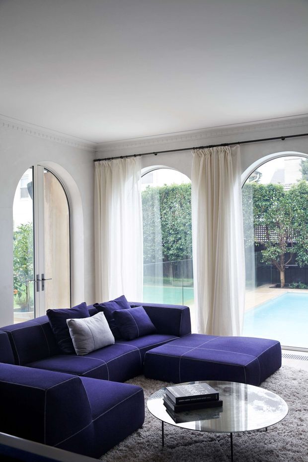 A bright, elegant living room with a deep navy modular sectional sofa and matching ottoman arranged around a round glass coffee table. Full-height arched windows and an arched glazed door are dressed with sheer cream curtains, providing views to a swimming pool and landscaped courtyard. Decorative ceiling cornices and pale walls add a refined traditional architectural character, while the plush rug and dark upholstery create a sophisticated, slightly dramatic atmosphere.