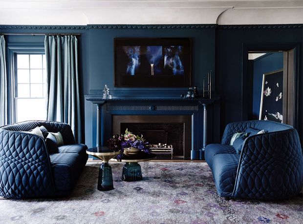 A richly styled formal living room with deep navy walls, matching quilted navy sofas and armchairs, a traditional fireplace with ornate mantel detailing, heavy blue curtains, white decorative ceiling moulding, and a patterned area rug. A framed artwork hangs above the fireplace, while a doorway opens to another dark-blue room.