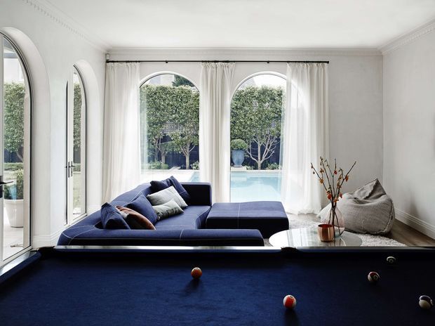 A bright, elegant living room with a deep navy sectional sofa and matching ottoman arranged beside a full-size dark pool table. Tall arched windows and glazed doors are dressed with sheer white curtains, overlooking a turquoise pool and landscaped courtyard. White walls, ornate ceiling cornices, and restrained furnishings create a refined, classic interior with strong navy-and-white contrast.