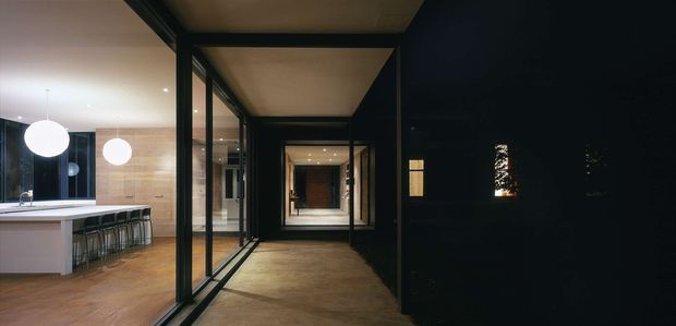 A contemporary interior view looking along a dark-framed glazed hallway or circulation spine. On the left, an open kitchen features a pale island with bar seating, timber cabinetry or wall panelling, and two spherical pendant lights. Full-height black-framed glazing and door frames create a series of layered thresholds, while warm timber-toned flooring and ceilings contrast with the dark walls and frames.