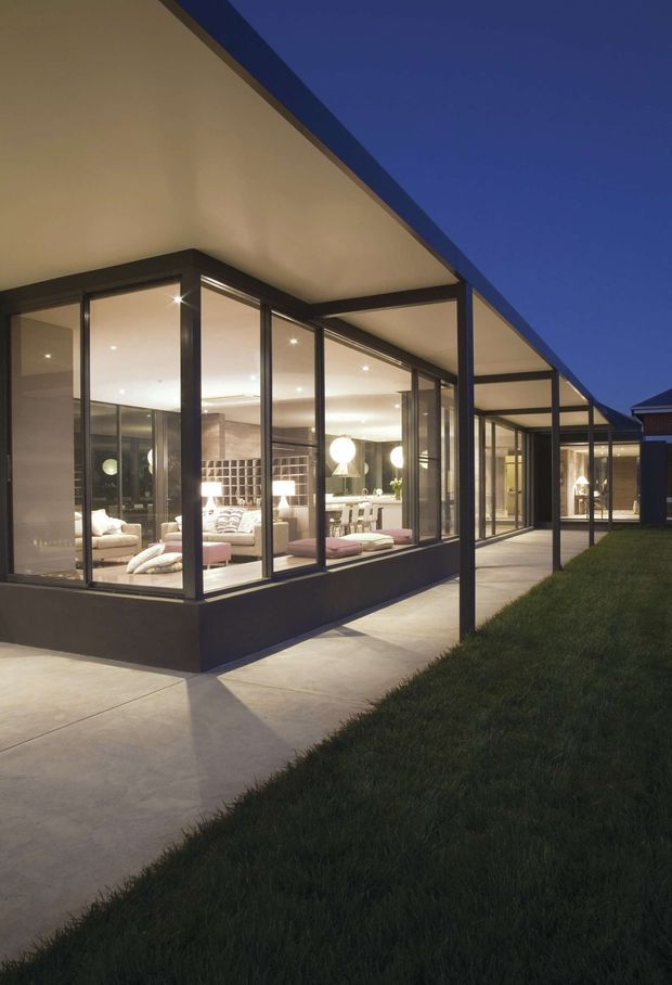A dusk exterior view of a modern single-storey pavilion-style home with extensive floor-to-ceiling glazing, dark slender metal frames, and a broad flat projecting roof. The glazed living and dining areas are illuminated inside, revealing contemporary furnishings, while a paved walkway and lawn surround the building.