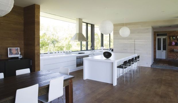 Bright open-plan kitchen and dining area with a large white island, integrated cabinetry, timber flooring, a long dark timber dining table, and a row of bar stools. Floor-to-ceiling glazing provides views to greenery, while sculptural pendant lights and a stainless-steel rangehood add contemporary detail. A hallway and adjoining spaces are visible in the background.