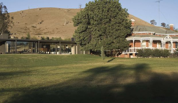 A wide exterior view of a rural property showing a historic ornate brick-and-verandah house beside a long, low contemporary glazed pavilion. The buildings sit on an expansive lawn with mature trees and dry rolling hills in the background. The composition highlights the contrast between heritage architecture and a modern glass extension.