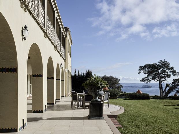 A grand Mediterranean-inspired waterfront terrace with a long colonnade of cream stucco arches, decorative patterned tile accents, ornate wall lanterns, and an upper balcony with lattice-style metalwork. The paved terrace contains an outdoor dining setting and large urn planter, opening onto a manicured lawn and expansive ocean views. The scene is bright and refined with classical resort-like detailing.