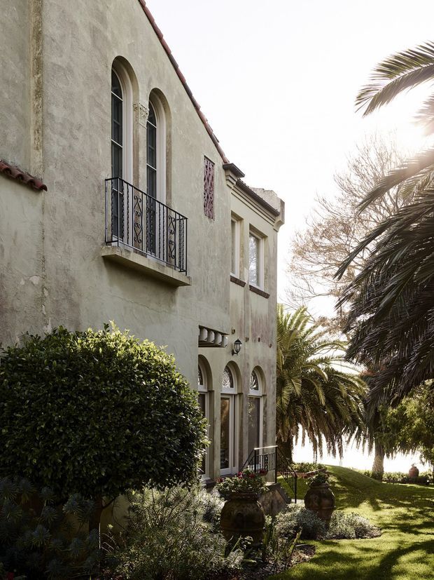 Exterior view of a Mediterranean-inspired stucco building with tall arched windows and doors, decorative wrought-iron balconies and railings, terracotta roof tiles, and ornamental wall detailing. The facade is set within a lush landscaped garden with mature palm trees, clipped hedges, lawn, and large decorative urns.