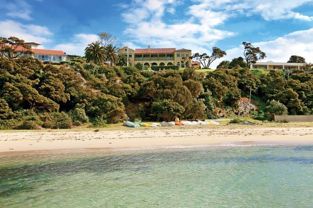 A wide coastal exterior view across clear shallow water and a sandy beach toward a landscaped, tree-covered residential shoreline. Several substantial homes sit above the beach, including a prominent warm-toned multi-level house with arched openings and terracotta-coloured roof tiles. Kayaks or small boats are stored along the beach. Bright blue sky and coastal vegetation create a relaxed seaside setting.
