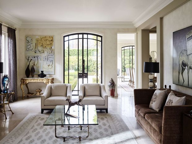 Elegant, traditionally detailed living room with a pair of upholstered armchairs, a brown sofa, glass coffee table, patterned area rug, ornate console table, decorative artwork and lamps. Tall black-framed glazed doors open toward a bright garden-facing area, while a wide opening leads to a dining space beyond. The room has ornate ceiling cornices, pale textured walls, polished light flooring and a refined, formal atmosphere.