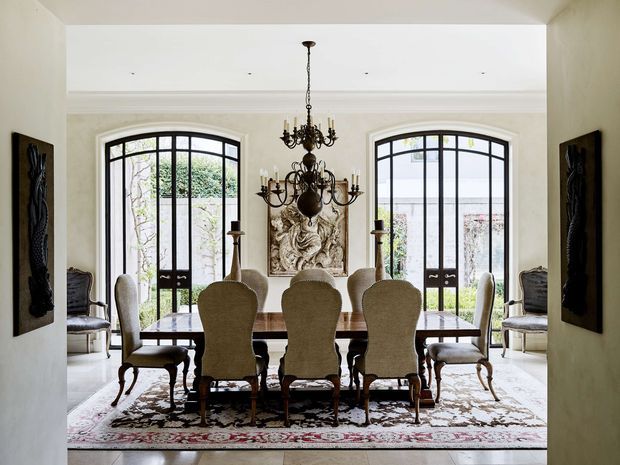 Formal dining room viewed through a wide opening, featuring a long dark timber dining table surrounded by upholstered high-back chairs, an ornate multi-arm chandelier, arched black-framed glazed doors, pale walls with decorative moulding, framed artwork, and a large patterned rug. The room has a refined traditional character with abundant natural light.