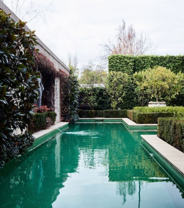 A formal landscaped courtyard dominated by a rectangular swimming pool with turquoise water. Pale stone or concrete coping and narrow pathways frame the pool, while dense clipped hedges, pleached trees, mature broadleaf planting, and a tall green hedge create a private garden enclosure. A simple rendered or concrete garden wall and an ornamental urn are visible in the background.
