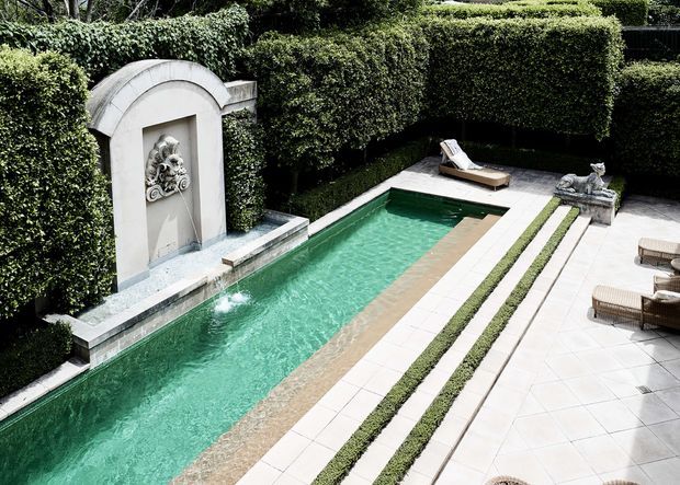 A formal rectangular swimming pool set within a meticulously manicured courtyard garden. The pool is bordered by pale stone paving and narrow clipped hedge strips, with tall sculpted hedges enclosing the space. A decorative arched stone fountain feature with a sculptural relief forms one end of the pool, while loungers and outdoor seating occupy the opposite terrace.