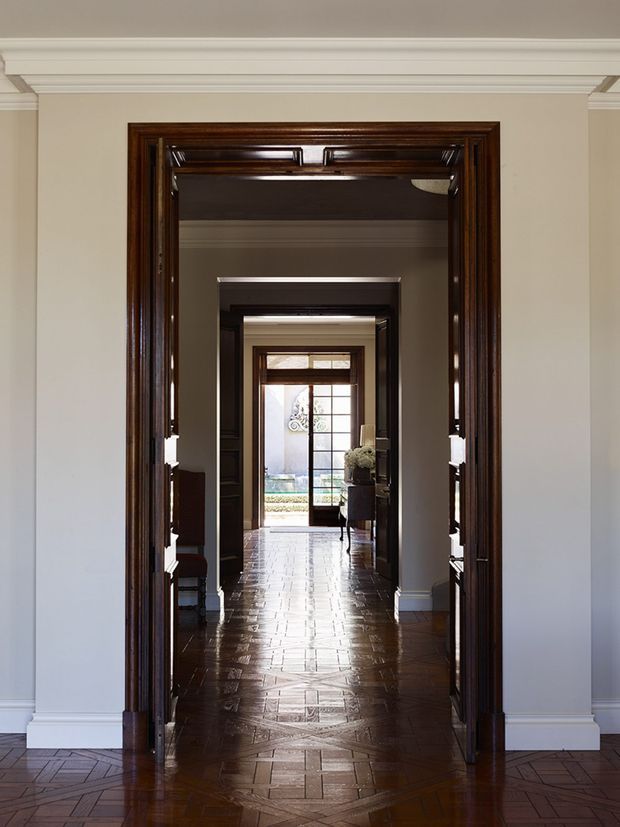 A symmetrical view through a sequence of richly stained dark timber doorways and trim, creating a long interior sightline toward a bright glazed rear door and courtyard beyond. The hallway has warm parquet timber flooring, cream-painted walls, white decorative cornices and skirting, and traditional architectural detailing.