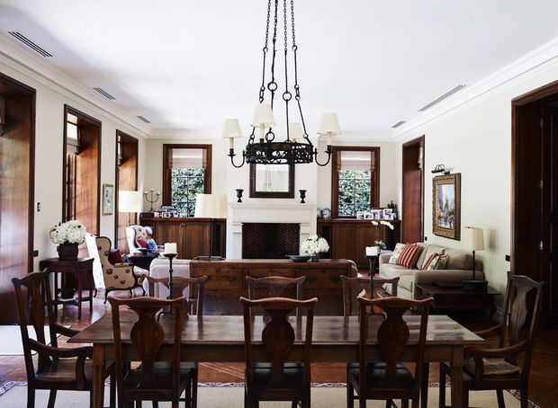 A formal, open-plan dining and living room with a large dark timber dining table and chairs in the foreground, facing a traditional fireplace and seating area. The room features rich timber wall and window detailing, white plaster walls and ceiling, a dark ornate suspended chandelier, classic upholstered furniture, and symmetrical built-in cabinetry around the fireplace. The overall presentation is refined and traditional with warm natural light.