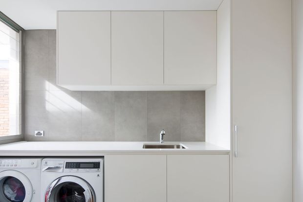 A bright, compact contemporary laundry with integrated white cabinetry, a long white benchtop, grey tiled splashback, stainless sink and tap, and front-loading washing machine and dryer beneath the counter. A large window brings in natural light.