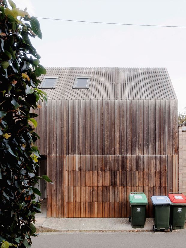 A narrow urban building facade clad entirely in vertically arranged weathered timber boards, including the pitched roof plane. Two small skylights sit high on the facade, while a large timber garage or entry door occupies the lower portion. Dense evergreen foliage frames the left edge, with wheelie bins and a paved street in the foreground.