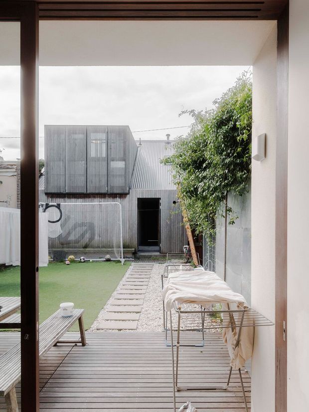 View from an interior doorway onto a compact landscaped courtyard and timber deck. The outdoor area includes artificial turf, a small soccer goal, stepping-stone path, gravel strip, leafy tree, and a covered outdoor drying rack or seating structure. The opening is framed in dark timber with a horizontal slatted detail overhead, while the courtyard boundary and adjacent outbuilding feature corrugated metal cladding.