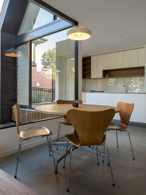 A bright, open-plan dining area adjoining a modern kitchen. A round timber dining table with sculptural mid-century-style wooden chairs sits beside full-height black-framed glazing, with views to a brick and timber courtyard. The kitchen features white flat-panel cabinetry, open timber shelving, a white subway-tile splashback, and a white island. Concrete flooring, dark window frames, and simple pendant lights create a clean contemporary interior with strong mid-century influences.