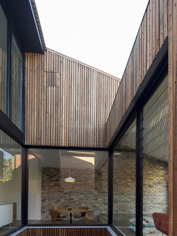 Interior view of a contemporary courtyard-like space framed by full-height glazing and dark metal window and door frames. Vertical timber cladding rises around the upper walls, while exposed warm-toned brickwork forms the lower-level walls. A compact dining area with a round table and chairs is visible beyond the glazing, with a pendant light overhead. The composition emphasises an enclosed courtyard, strong vertical lines, and abundant natural light.