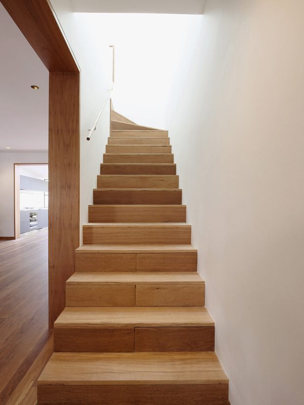 A modern interior stairway viewed from the lower level, featuring broad solid timber treads and risers with visible grain, set against smooth white walls. A slim minimalist handrail follows the stair edge, while a timber-clad structural opening frames a glimpse of the adjacent hallway and room. The stair rises toward a bright upper landing.