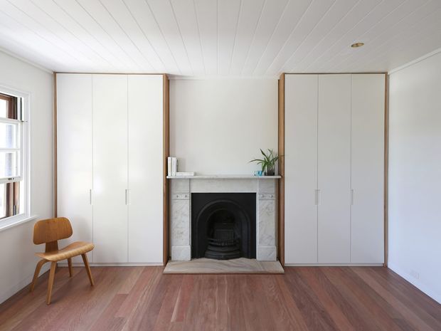 Bright, restrained bedroom interior with tall white built-in wardrobes framing a central traditional fireplace with a dark arched insert and pale stone surround. The room has warm timber flooring, white painted walls and a white-lined ceiling, with a simple timber chair by the window and minimal decor. Natural light enters from the side window, creating a calm, clean and airy atmosphere.