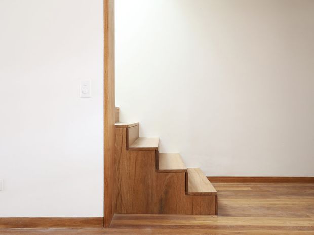 Minimal interior view of a compact timber staircase with alternating stepped storage-like side panels, rising beside a white plaster wall. The staircase and surrounding floor feature warm natural wood, with simple white walls and restrained detailing.