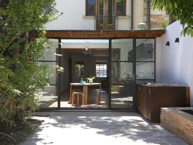 A leafy urban courtyard viewed from outdoors, showing a contemporary two-storey residence with a glazed ground-floor extension. Slim dark-framed sliding glass doors open into a compact dining area, while a timber-framed roof or canopy spans the facade. White rendered walls, exposed brickwork, timber joinery, and a dark built-in outdoor cabinet line the courtyard. Dappled sunlight and mature planting create a sheltered connection between the interior and exterior.