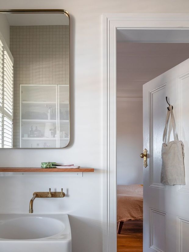A refined bathroom vanity area with a white basin, wall-mounted brass tapware, timber shelf, and a large rounded-corner mirror. Small square wall tiles and white plantation shutters are reflected in the mirror. A paneled door opens directly to a bedroom with a visible bed and timber floor, establishing the bathroom as an ensuite.