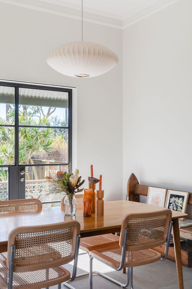 A bright dining room with a rectangular timber dining table and woven cane dining chairs. The room has pale walls with decorative ceiling cornices, a large black-framed glazed door or window, framed artwork leaning against the wall, and a sculptural white pendant light. Natural greenery is visible outside, contributing to an airy, warm, mid-century-inspired interior.