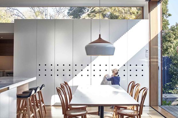 Bright contemporary open-plan dining area with a long white dining table surrounded by curved timber chairs. A large sculptural pendant light hangs above, while a perforated white partition wall and sculptural staircase create a strong architectural backdrop. Floor-to-ceiling glazing and timber framing connect the interior to leafy surroundings, with a kitchen and island visible to the left.