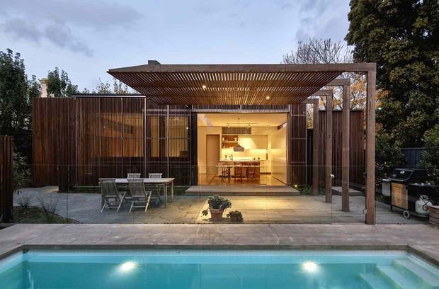 Exterior view of a contemporary single-storey pavilion-style home opening onto a tiled terrace and swimming pool. The facade combines dark vertical timber screening with large glazed sliding doors, while a broad timber pergola with slatted roof provides shade over the outdoor entertaining area. A warmly lit kitchen and dining space is visible through the glazing, with outdoor furniture and a built-in barbecue beside the terrace.