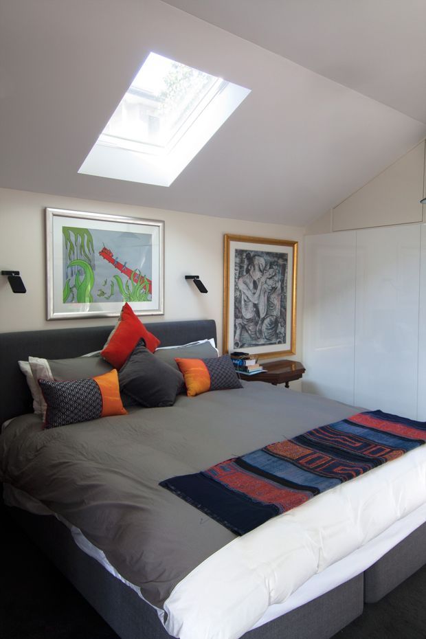A compact, contemporary bedroom viewed from near the foot of a large upholstered bed. The room has a sloped white ceiling with a prominent rectangular skylight, built-in white wardrobe cabinetry along the right wall, dark carpet, and a dark charcoal upholstered bedhead. Layered grey bedding is accented with vivid red-orange and patterned cushions, while framed artwork and small wall-mounted reading lights decorate the walls.