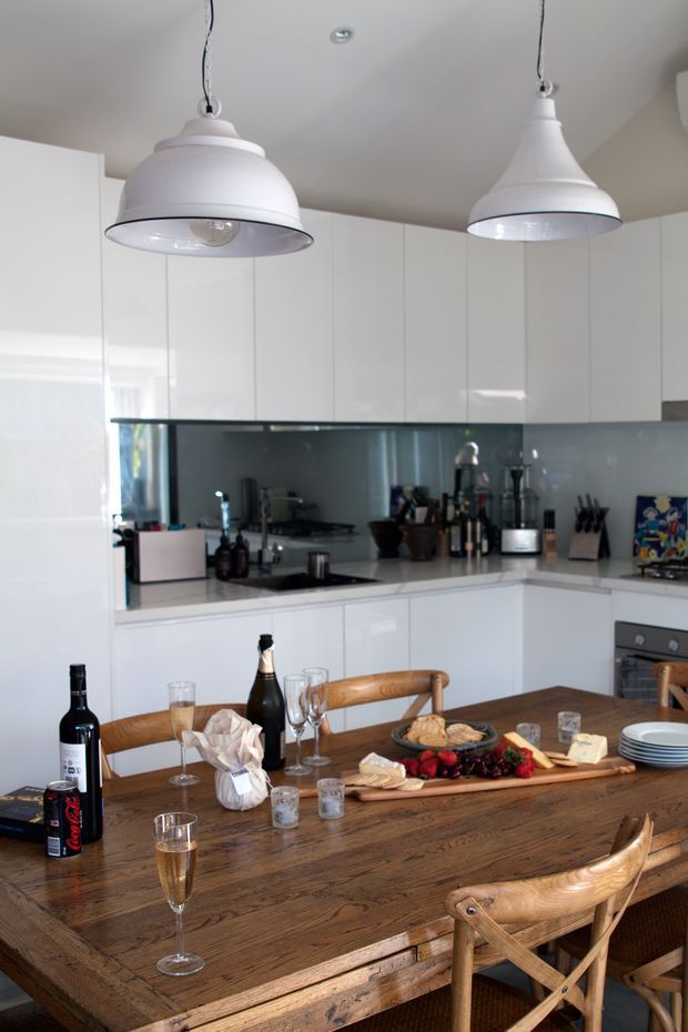 Bright contemporary kitchen and dining area with glossy white full-height cabinetry, a dark reflective splashback, white benchtops, and two oversized white pendant lights. A large rustic timber dining table with cross-back wooden chairs occupies the foreground and is set with wine, glasses, plates, and a grazing platter.