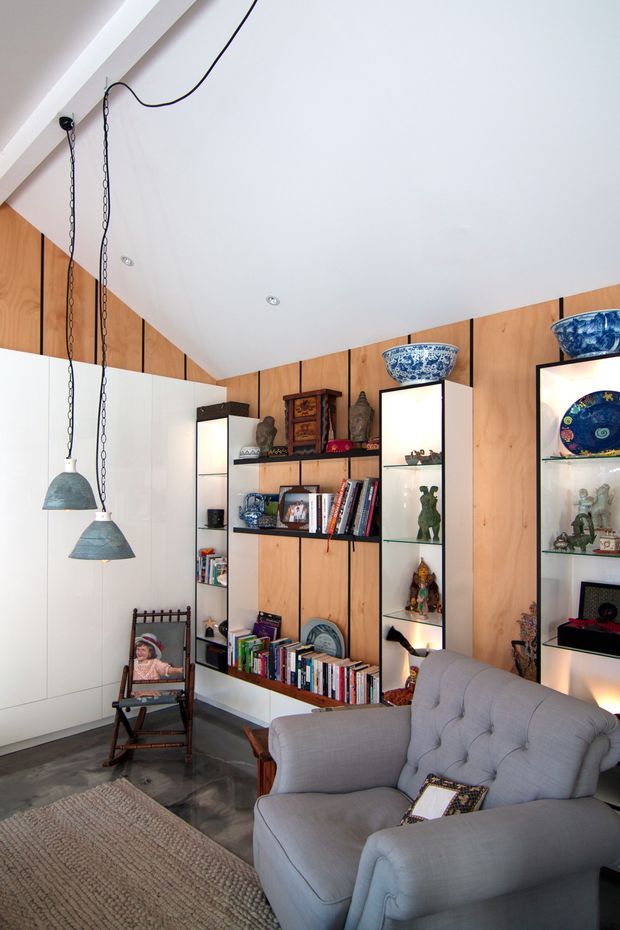 A bright, eclectic living room with a sloped white ceiling, exposed timber wall panelling, built-in open shelving, and illuminated display niches filled with books, ceramics, and decorative objects. A large grey upholstered sofa, rocking chair, woven rug, and hanging pendant lights occupy the room. The space has a strong mid-century-inspired built-in joinery aesthetic with personal, collected décor.