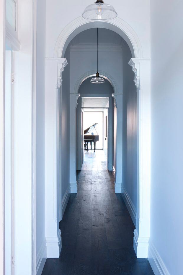 A long, narrow residential hallway with repeated arched openings, ornate white classical mouldings and columns, pale blue-grey walls, dark timber flooring, and pendant lights aligned along the corridor. Bright daylight enters from the far end, where a grand piano is partially visible in an adjoining room.
