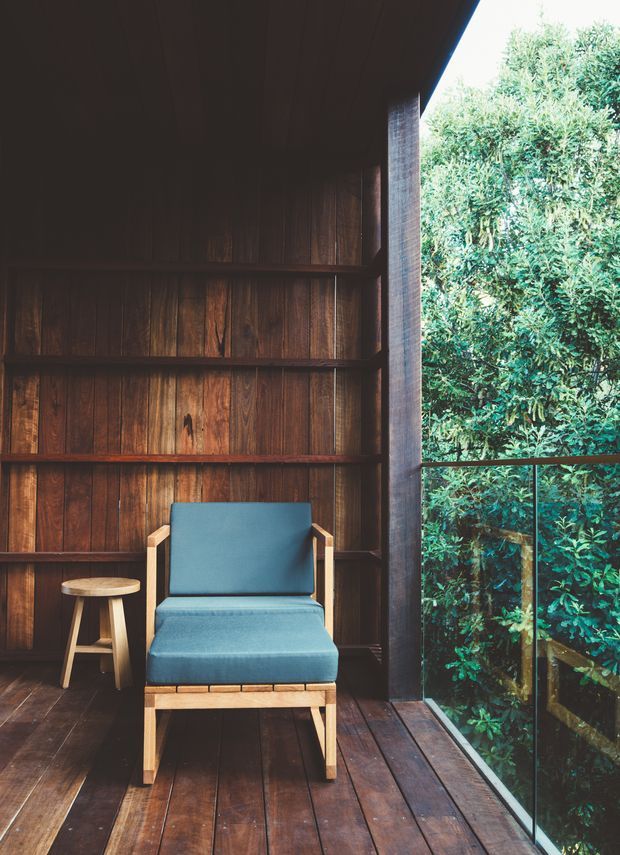 A compact, quiet timber-lined interior or enclosed terrace with dark vertical wood wall panelling, matching timber floorboards, and a large floor-to-ceiling glazed opening overlooking dense greenery. A blue upholstered lounge chair with light timber arms and legs sits beside a small round wooden stool. The space has strong natural-material character, high contrast between the dark timber and bright foliage, and a calm indoor-outdoor connection.