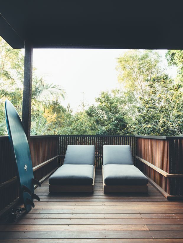 A sheltered elevated timber deck or balcony surrounded by dense greenery, with two cushioned lounge chairs and a vertically stored surfboard. Dark timber posts, balustrades, and overhead roof structure frame the open outlook.