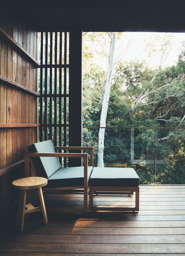 A tranquil timber-lined outdoor room or enclosed balcony with a timber deck, built-in vertical slatted screening, full-height clear glazing, and a lounge chair with matching ottoman. The space overlooks dense native-looking greenery and is styled with warm natural timber, dark framing, and muted pale upholstery.