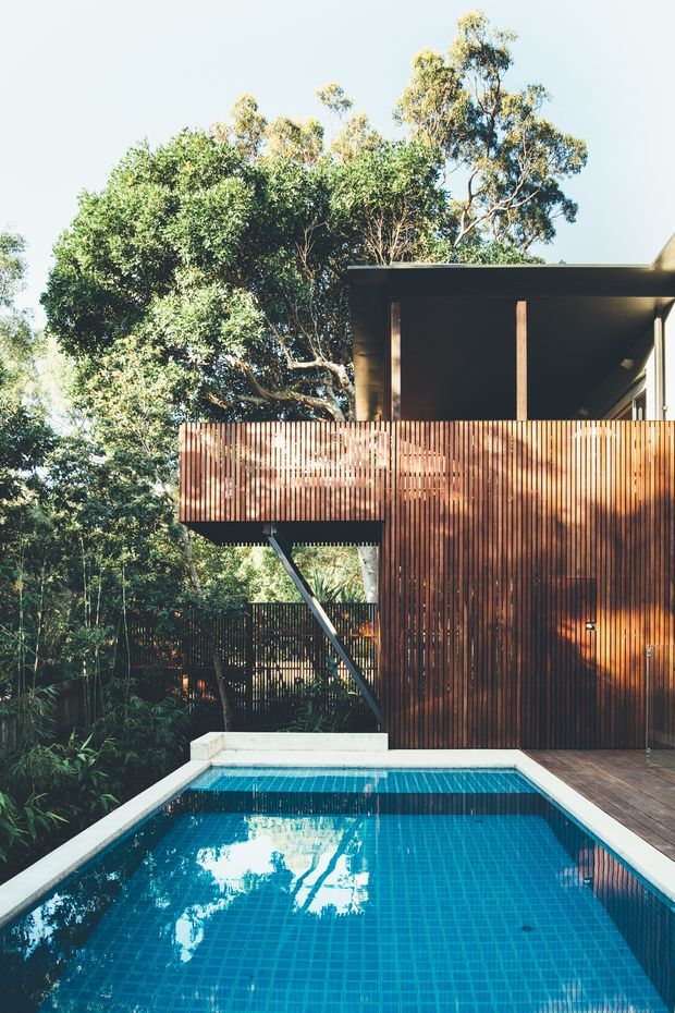 Exterior view of a contemporary multi-level home set among dense vegetation, with a projecting upper deck enclosed by vertical timber slats, dark flat-roofed pavilion structure, and a blue tiled swimming pool in the foreground.