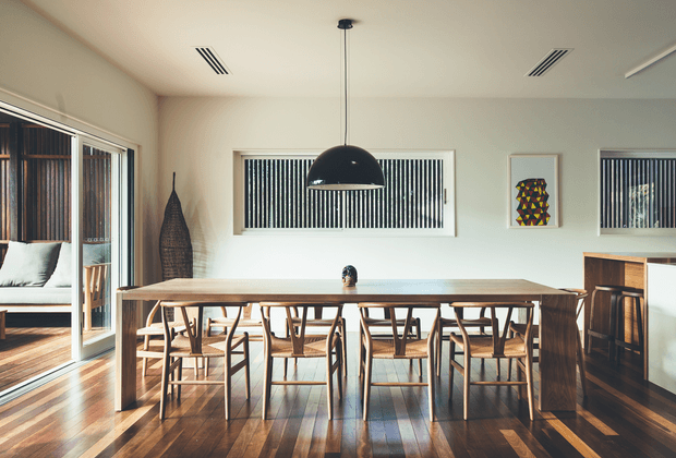 Bright, warm-toned dining room with a long solid timber dining table surrounded by timber chairs, polished timber flooring, and a large black dome pendant light. White walls feature horizontal windows with dark vertical blinds, while glazing opens to a timber-floored outdoor or adjoining area. A compact built-in timber bar or console occupies the right side, with a framed geometric artwork above it.