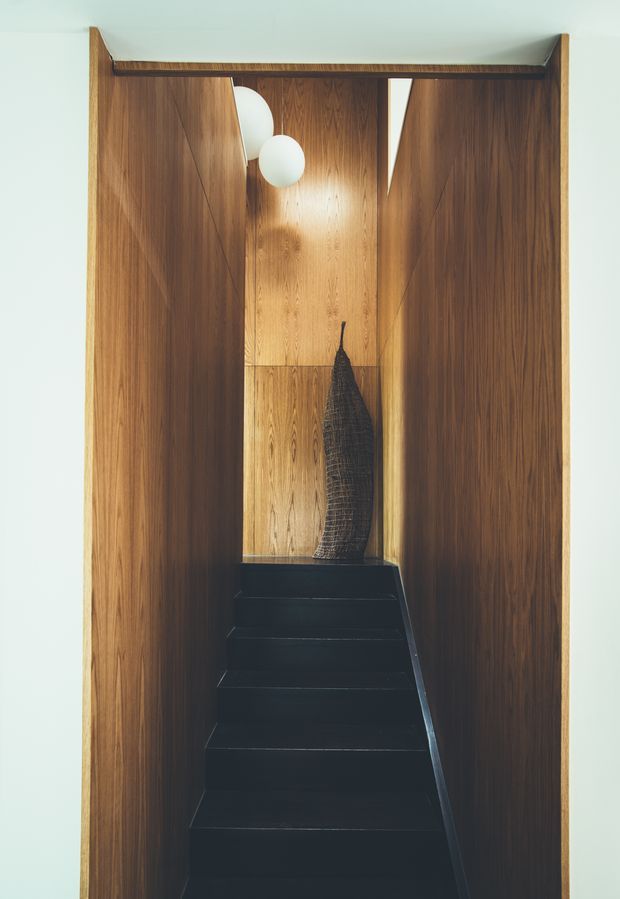 A narrow, vertically framed stairway enclosed by rich dark timber wall panelling and matching overhead trim. The stairs are finished in black, leading to a small landing with a sculptural dark vase and warm timber panel backdrop. Two white spherical pendant lights hang near the upper portion, adding a soft modern accent. The composition is warm, refined, and highly architectural, with strong symmetry and concealed detailing.