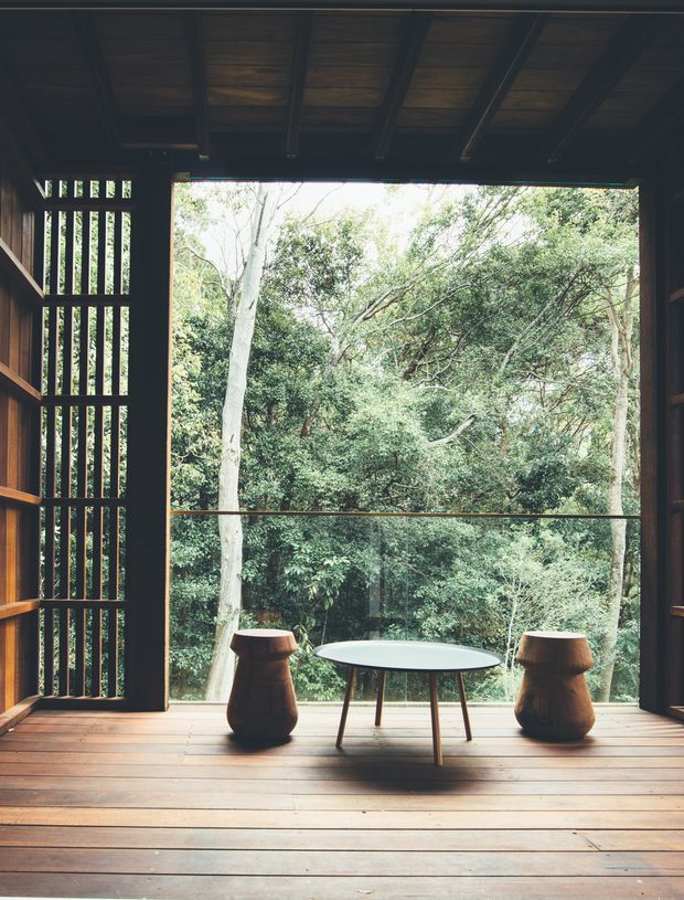 A dark timber-lined interior opens onto a lush wooded landscape through a large glazed opening. The room features exposed timber roof beams, vertical timber slat screening, wide timber floorboards, and a simple round table with two sculptural wooden stools. The composition is quiet and nature-focused, with strong indoor-outdoor connection and abundant natural light beyond the opening.