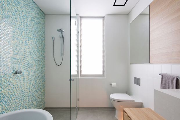Contemporary bathroom with a frameless glass shower enclosure, blue-green mosaic feature wall, white wall surfaces, grey floor tiles, wall-hung toilet, floating timber-toned vanity cabinetry, mirrored storage, and a narrow window providing natural light.