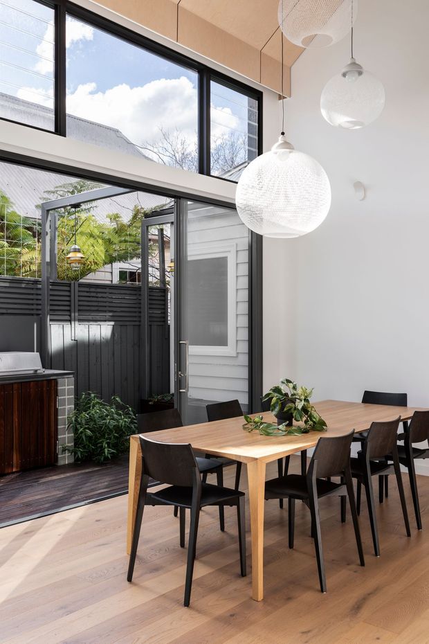 Bright contemporary dining room with a large light-wood dining table and dark upholstered chairs, positioned beside full-height black-framed glazing and a glazed door opening to a compact outdoor courtyard. Three white perforated pendant lights hang from a high ceiling, while pale timber flooring and light walls create an airy, refined interior. A glimpse of an outdoor kitchen or barbecue area is visible beyond the glazing.