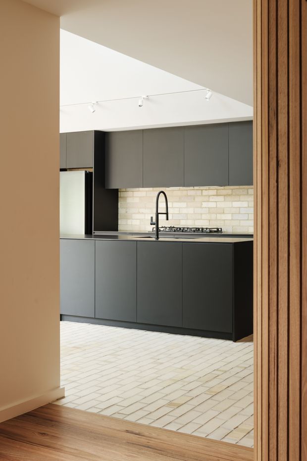 A contemporary kitchen viewed through a timber-framed opening. The room features dark charcoal flat-panel cabinetry, a pale brick or rectangular-tile splashback, a stainless-steel benchtop, integrated appliances, a black gooseneck tap and gas cooktop. Pale rectangular floor tiles transition to warm timber flooring in the foreground. A high white ceiling with track lighting creates a bright, clean space.