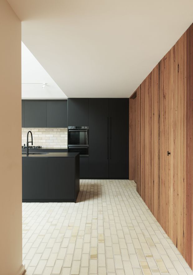 A contemporary kitchen viewed from an adjoining space, featuring matte black full-height cabinetry, a black island with integrated cooktop and tall curved faucet, pale brick-pattern tiled flooring, a light brick splashback, and a prominent vertical timber slat wall with concealed storage. The room has a dramatic angled ceiling and a restrained palette of black, warm timber, cream and pale beige.