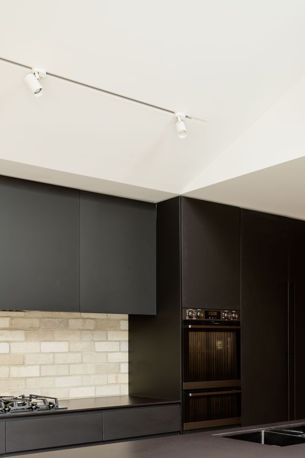 A modern minimalist kitchen with full-height matte dark cabinetry, integrated double ovens, a dark benchtop, and a pale exposed-brick splashback. A gas cooktop and sink are visible along the lower cabinetry, while white track lighting illuminates the clean, angular space.