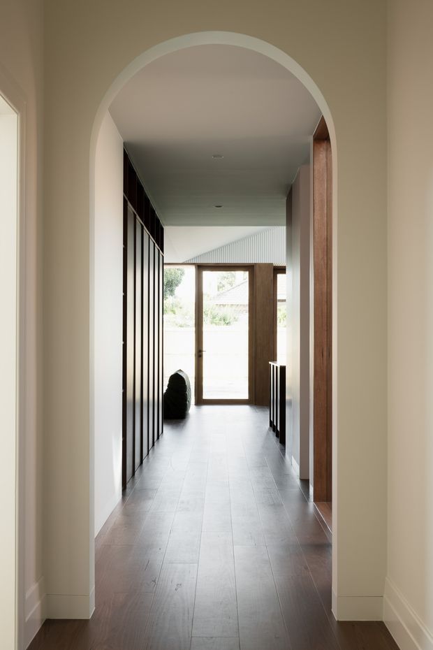 A refined residential hallway viewed through a large rounded archway. The corridor has dark timber flooring, full-height dark timber cabinetry or wall panelling on the left, warm timber door and trim elements on the right, pale painted walls and ceiling, and a glazed timber door at the far end opening toward an outdoor area. The composition is calm and minimal with strong axial perspective and natural light.