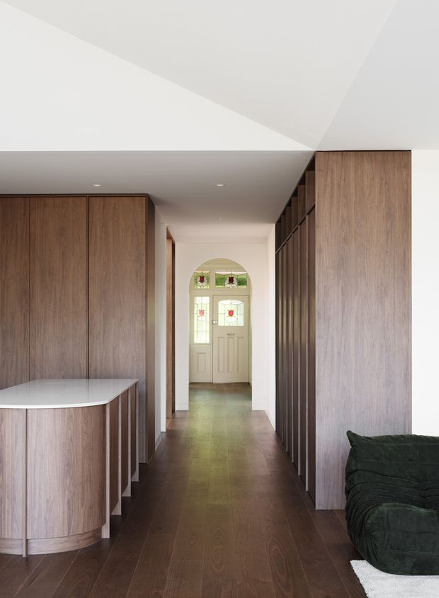 Interior view down a narrow contemporary hallway with extensive dark timber built-in cabinetry and wall panelling. A curved timber peninsula with a light benchtop occupies the left foreground, while a dark green sofa is partially visible on the right. The hall leads to a traditional glazed front door with decorative coloured glass, creating a contrast between the modern timber-lined interior and heritage-style entry. White ceilings and walls, recessed downlights, and dark timber flooring complete the space.