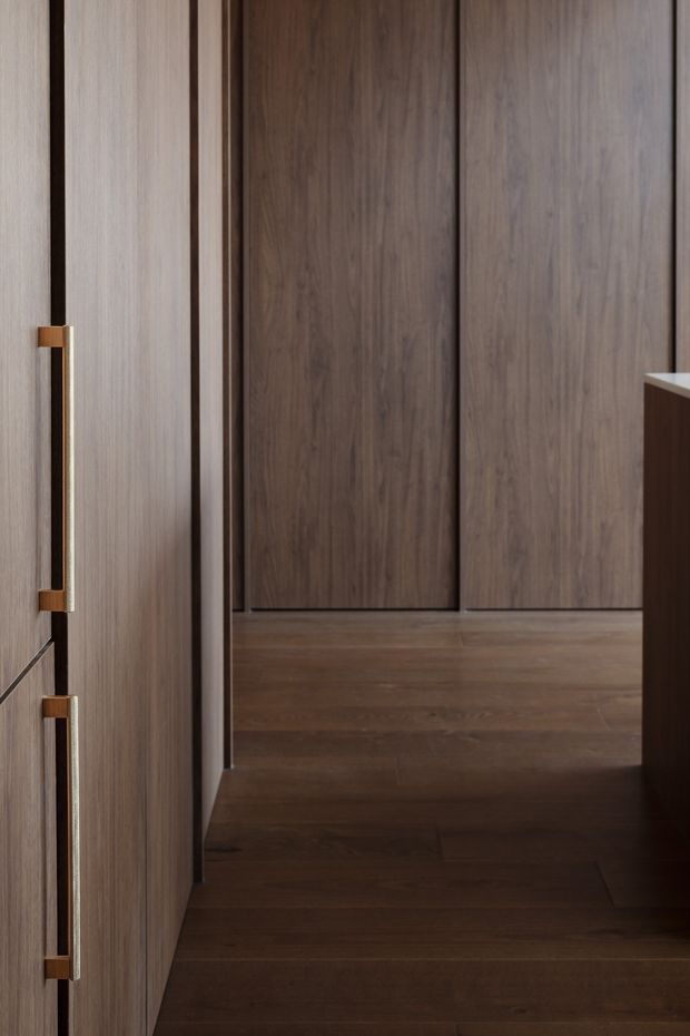 A close-up view of a dark timber-lined interior passage or built-in storage area. Floor-to-ceiling wood panels and cabinetry create a continuous vertical rhythm, with long slim metal and timber handles visible on the left. The space is minimally styled, warm, and dimly lit, with a small glimpse of a pale benchtop and dark joinery at the right edge.
