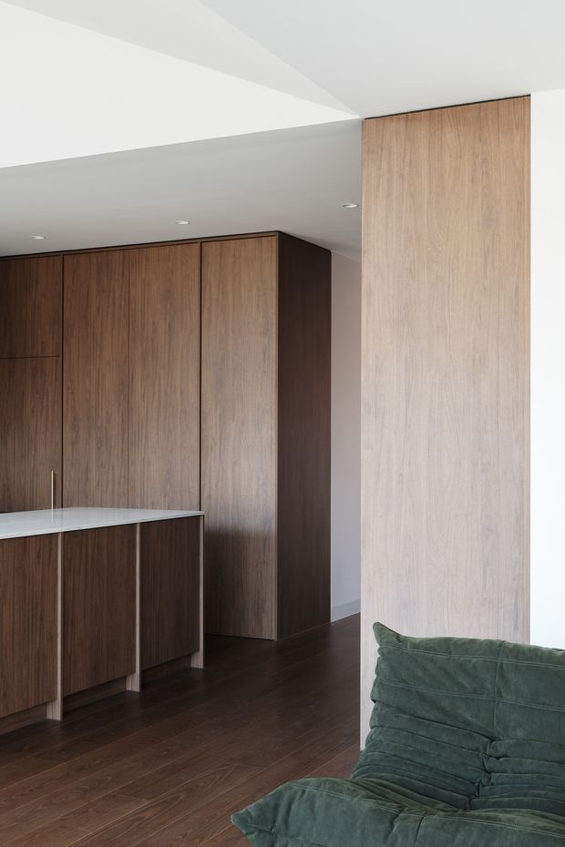 A refined open-plan kitchen and living area with dark timber cabinetry, a pale stone island benchtop, and matching full-height timber wall panelling. The room has dark timber flooring, a white ceiling with recessed downlights and a sculptural angled bulkhead, while a plush deep green lounge chair occupies the foreground. The overall composition is clean-lined, understated, and contemporary.