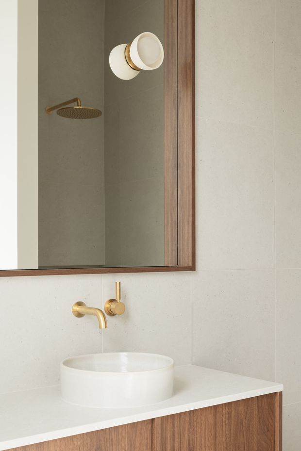 A close-up contemporary bathroom vanity with a round white vessel basin on a white benchtop, warm brushed-gold wall-mounted tapware, and a timber-veneer vanity cabinet. Large light beige-grey wall tiles and a framed timber mirror dominate the scene; the mirror reflects a matching gold showerhead and wall light.